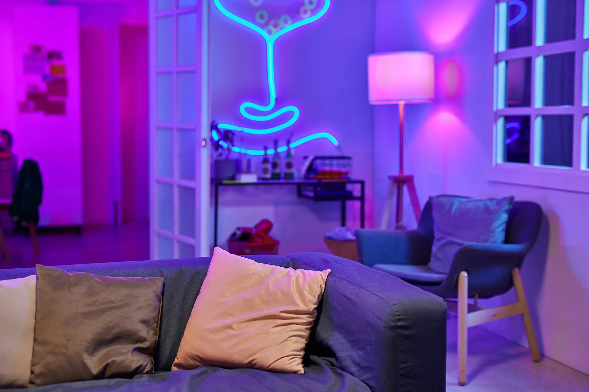 A cozy living room with gray and gold pillows on a couch. A neon light in the shape of a face decorates the wall above a shelf with books and bottles. A gray armchair and a lit floor lamp stand next to a window, illuminated by pink and blue lights.